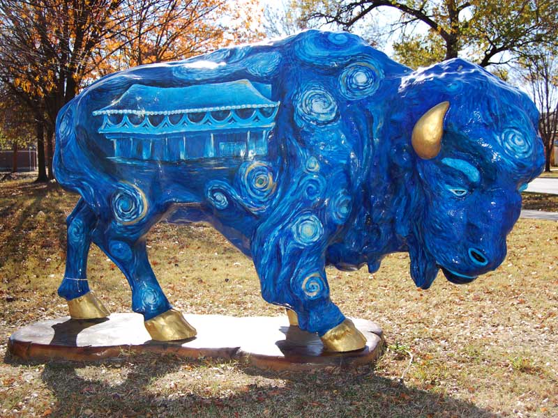 Painted buffalo sculpture with inspired by "A Starry Night." Depicts Bartlesville Community Center and downtown Bartlesville skyline with blue swirling sky.